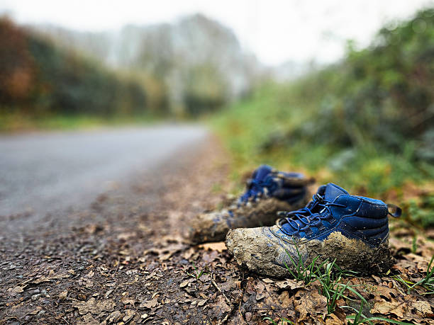 Low angle image depicting a pair of hiking boots, caked in mud, left abandoned at the side of the road. Focus on the boots with the receding perspective of the road defocused beyond.