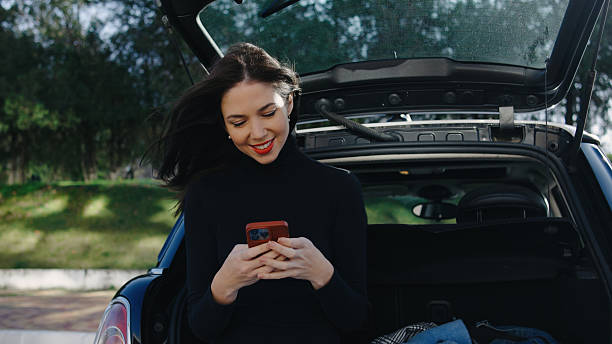 istock Young woman smiling while using smartphone outdoors next to open car trunk in sunny weather 2252363901