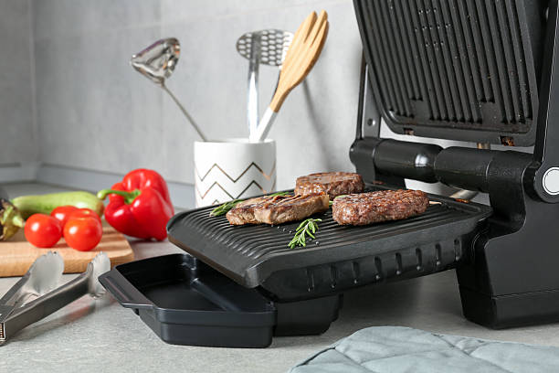 istock Open electric grill with beef steak, cutlets and rosemary, vegetables and cookware on light grey table, closeup 2252364399
