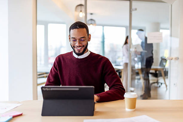 Smiling Man Working on Tablet at Modern Office Desk with Coffee Cup in Bright, Open Workspace