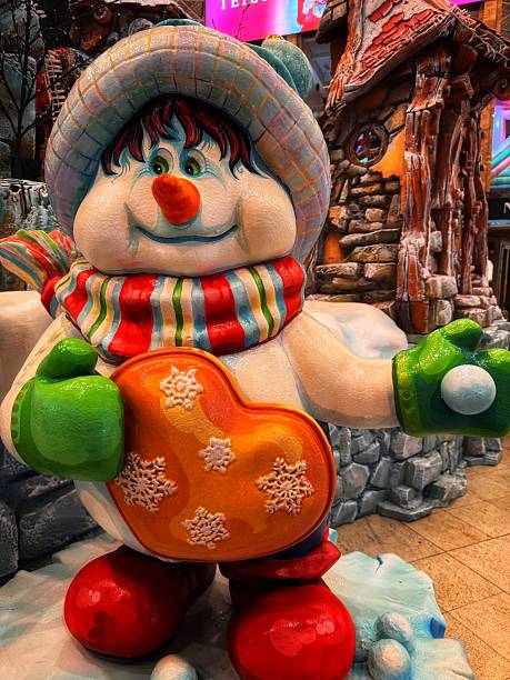 A brightly colored snowman figure stands at the center of a festive winter scene, holding a heart-shaped cookie decorated with delicate snowflakes. Dressed in a striped scarf, green mittens, and a cozy hat, the snowman’s cheerful expression brings a playful and welcoming spirit to the display. The detailed backdrop of snow-covered ground and rustic buildings enhances the storybook atmosphere, evoking warmth, joy, and the simple pleasures of the holiday season.