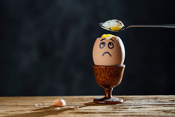 istock Metal spoon breaking boiled egg with sad face in egg cup on wooden table, closeup. Scared egg looking at spoon 2252370432