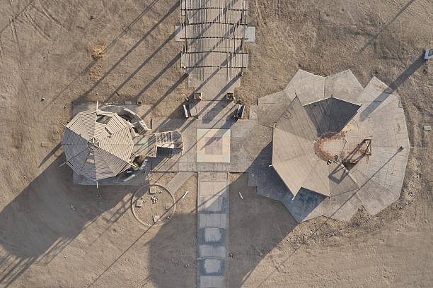Aerial top view of modern geometric architectural structures connected by walkways in an arid desert environment. Abstract symmetry, concrete forms, and long shadows create a minimal and graphic composition with no people present.