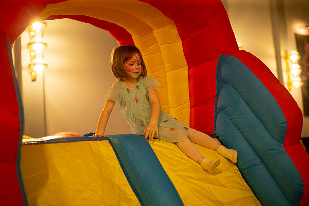 Young Caucasian Girl On Inflatable Slide Smiling Under Rainbow Arch At Indoor Party Venue, Warm Lighting And Soft Focus. Image Illustrates Carefree