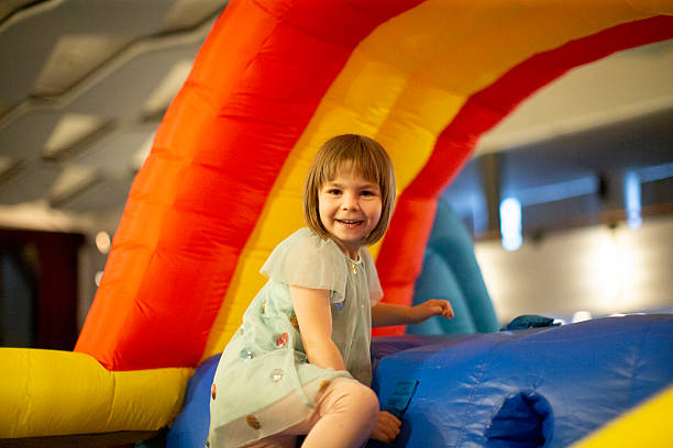 Young Caucasian Girl Smiling At Camera In Closeup Portrait With Rainbow Inflatable Backdrop And Warm Lighting, Candid Expression And Bright Eyes.