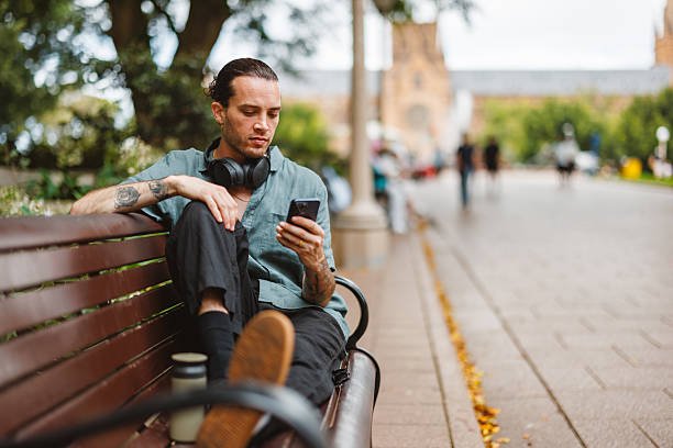 Mid adult Caucasian non-binary person sitting comfortably on a park bench in Sydney, focused on their smartphone. Surrounded by greenery and a peaceful atmosphere, they appear content and engaged, enjoying a leisurely moment in the vibrant outdoor setting.
