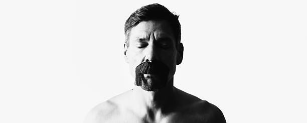 A man with closed eyes wears an artificial mustache. The minimalist black-and-white scene addresses contemporary questions of masculinity, self-image, and identity as a consciously shaped state between authenticity and performance.