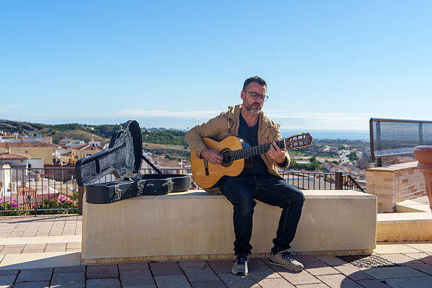 Man practicing guitar on a terrace overlooking a cityscape and the ocean. He is wearing glasses, a jacket, and jeans