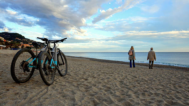 Mature couple park bicycles on empty beach at sunset, Liguria