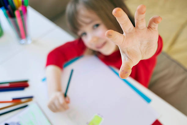 Child sits at table with colored pencils and paper, extending hand playfully during creative moment.