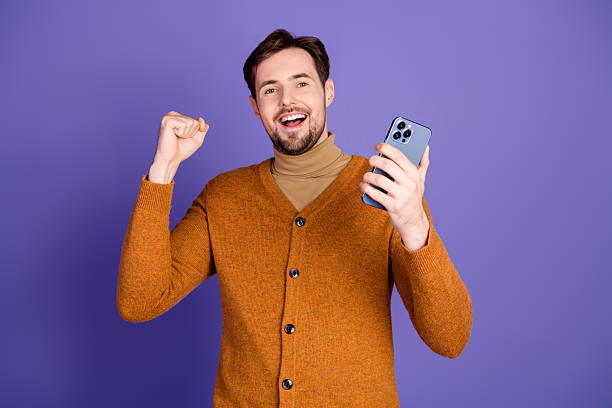istock Young man with orange cardigan celebrates while using a smartphone against a purple background in a casual stylish lifestyle image 2252415919