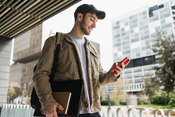 istock Young man walking in city while checking smartphone and carrying laptop 2252416163