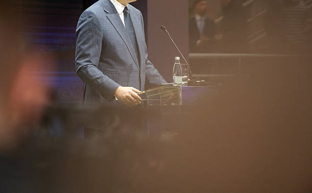 A political leader stands steadily at a podium, delivers a powerful speech, holding onto a podium with a folder of materials and a bottle of water, copy space