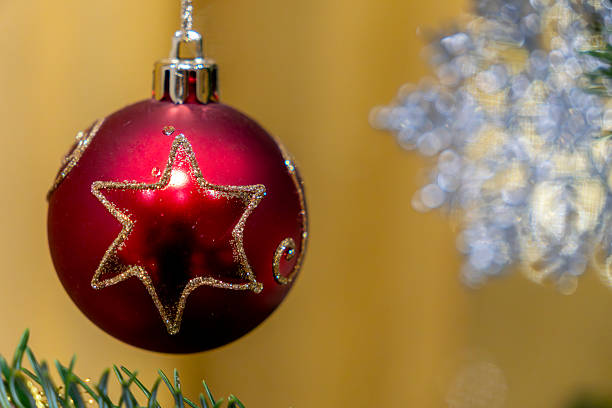 A red ornament with a star design is hanging from a pine tree branch. The background is blurred, creating a focus on the ornament. The scene shows festive decorations during the Christmas season.
