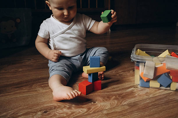 Baby sits on a wooden floor, playfully stacking colorful toy blocks from a nearby container, displaying concentration and curiosity. The scene captures a moment of early development and learning.