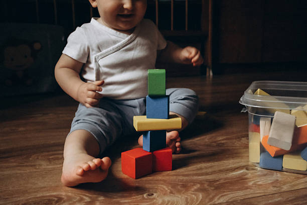istock Baby sits on a wooden floor, playfully stacking colorful toy blocks from a nearby container, displaying concentration and curiosity. The scene captures a moment of early development and learning. 2252425200