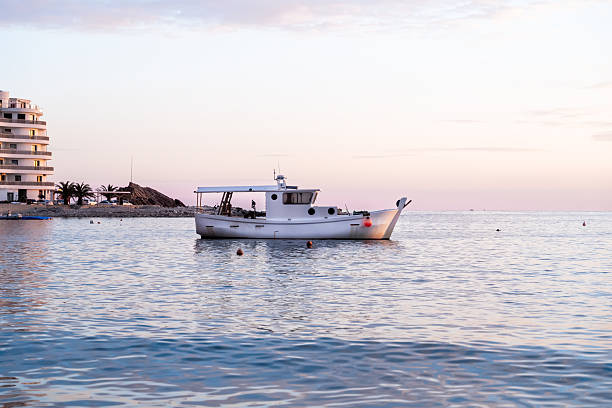 A serene white fishing boat drifts on calm waters during a beautiful pastel-hued sunset. The soft reflections of the sky on the water create a tranquil atmosphere, while a distant coastal landscape with palm trees and modern buildings adds depth to the scene.