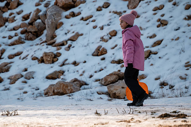 Child wearing winter clothes and pink beanie walking up a snowy mountain carrying an orange sled. Winter outdoors activity