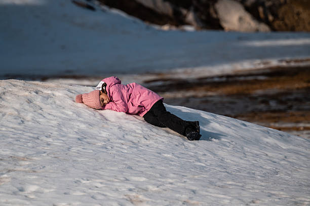 Child wearing a pink jacket and knitted hat resting face down on the snow, feeling cold and exhausted during winter