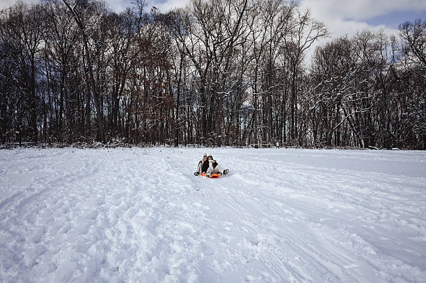 Two person sledding on an orange plastic sled down a snowy hill with bare trees in the background