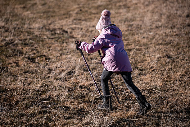 Young child wearing winter clothing and using trekking poles, walking uphill on dry grass outdoor, being active