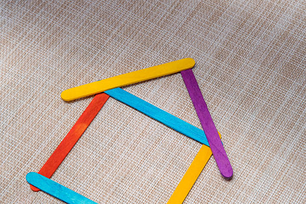 istock Multicolored craft sticks are arranged in an unstable outline of a basic house shape. 2252440417