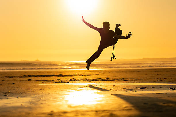 Energetic man jumping in a capoeira move, silhouetted against a golden beach sunset
