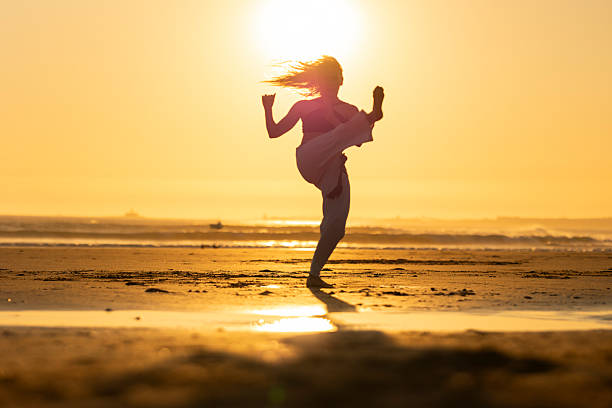 Woman performing a high kick on a golden beach at sunset, showing strength and freedom