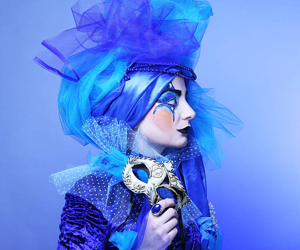Young woman with creative visage and costume holding mask over blue background.