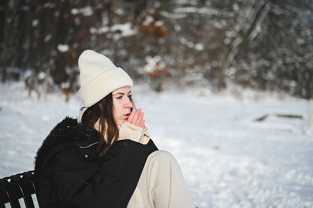 Young woman sitting on a park bench warming her cold hands in a snowy winter landscape