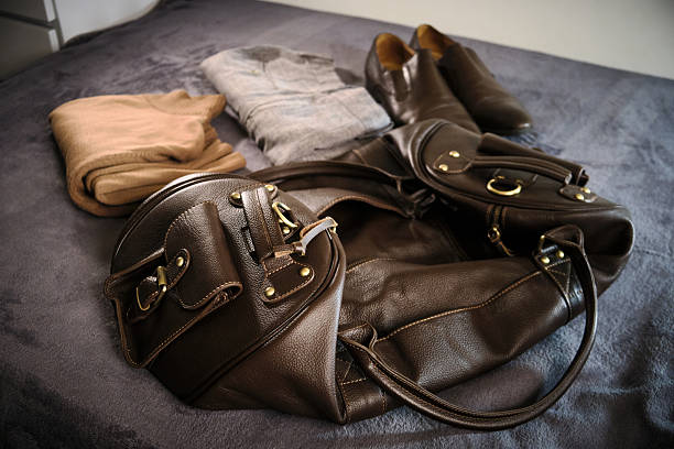 Brown leather travel bag lies open on bed alongside neatly folded clothing items, including sweater, pants and shoes. Packing for trip, organization, and readiness for travel