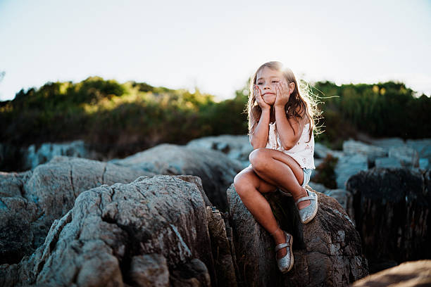 A five-year-old Eurasian girl sits on coastal rocks and makes a silly face while visiting the New England shoreline during a summer family road trip.