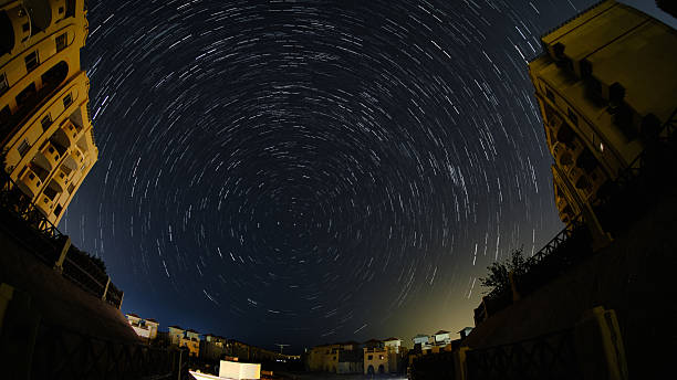 Long-exposure star trails swirl across night sky above residential buildings. Fisheye ultra-wide shot shows circular motion perspective of rotating stars