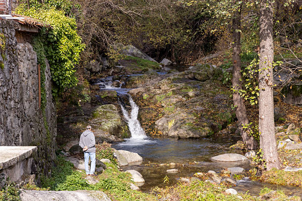 Cuevas del Valle, Avila, Spain, December 10, 2025: A man is sitting by a stream with a waterfall in the background. The scene is peaceful and serene, with the sound of the water and the lush greenery surrounding the man