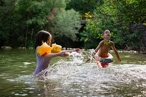 Two children playing and splashing water in a natural river surrounded by green trees on a warm summer day