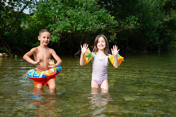 Two cute little girls sitting on a wooden platform by the river or lake dipping their feet in the water on warm summer day