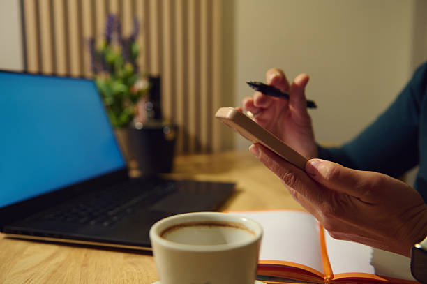 A modern cafe workspace shows a woman multitasking on a smartphone while a laptop, coffee, and open notebook support productive, relaxed work in a stylish setting.