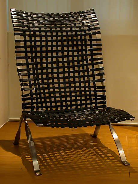 View of a chair with braided leather upholstery held in place by chrome, beige floor and white curtain in the background