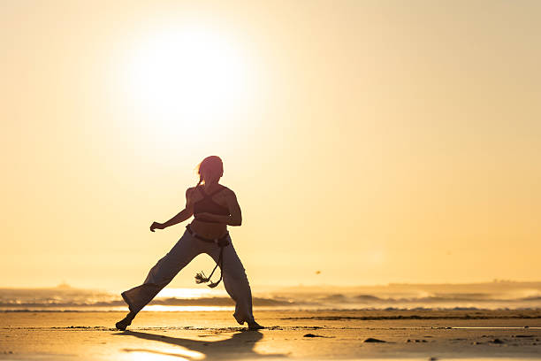 Woman performing capoeira movement on sandy beach silhouette during golden hour