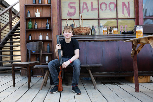 Boy holding long gun on bench. Young man in cowboy era saloon. Playful western themed scene for kids entertainment.