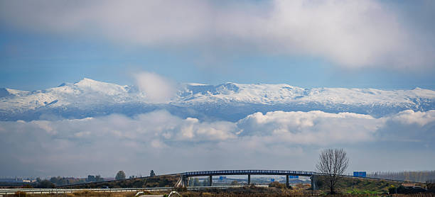 Snowy sierra nevada mountains forming a dramatic backdrop above a modern highway bridge and low-lying clouds across the fertile plains of vega de granada, spain