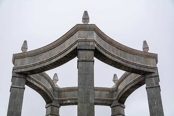 Low Angle View Of A Granite Monument Structure With Curved Beams And Central Column Against Overcast Sky, Strong Geometric Architecture For Heritage And Design Themes.