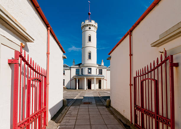 A white lighthouse tower stands at the end of a paved courtyard, flanked by red iron gates, under a clear blue sky. A picturesque coastal landmark with architectural charm.