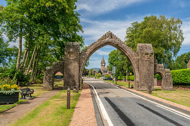 A weathered stone arch stands over a rural road in Edzell, Scotland, framed by lush trees and clear skies, evoking heritage, timeless village charm, and scenic travel appeal.