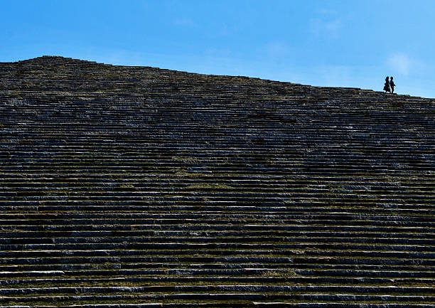 Stone steps form a broad, ascending structure under a clear blue sky. Two silhouetted figures stand near the top, highlighting the scale.