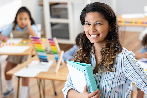 istock Smiling teacher holds a binder in a classroom setting with students in the background. 2252536678