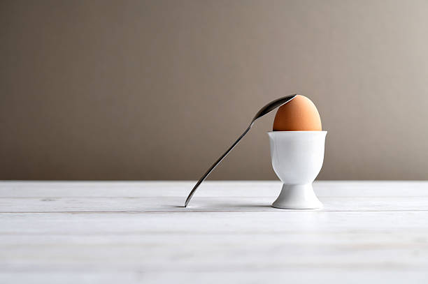 brown egg in a white ceramic egg cup and spoon on a light wooden surface. Clean composition, simple food concept and copy space for text.