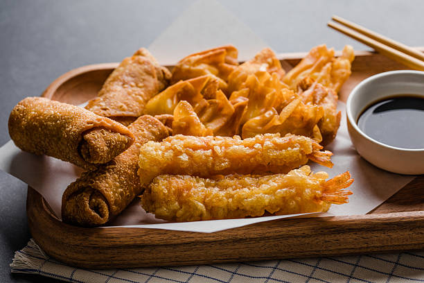 Asian appetizers including a crispy egg roll, fried cream cheese wontons, and a pork dumpling (potsticker) served with soy sauce on a dark textured background.
