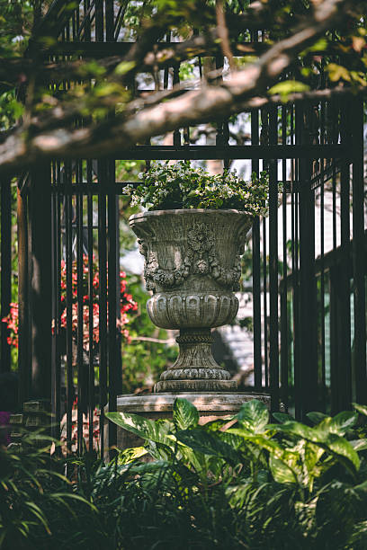 Elegant Roman-style stone planter with small white flowers inside a wrought iron structure. Surrounded by green foliage and dappled sunlight in a peaceful garden setting.