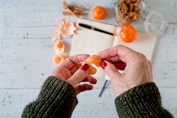 Close-up winter lifestyle flat lay showing female human hands peeling a fresh tangerine above an open diary, cinnamon sticks, and seasonal elements on a white wooden background. The natural action and soft tones convey warmth, comfort, and mindful winter living, with copy space for lifestyle or editorial concepts.